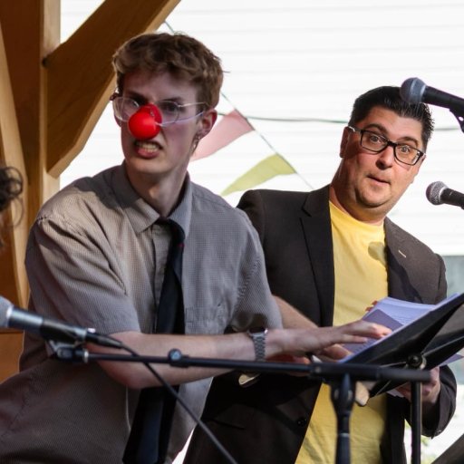 Archive photo of a Round Back performance. Two men standing at microphones at microphones look toward a seated man. One of the men is wearing a red clown nose.