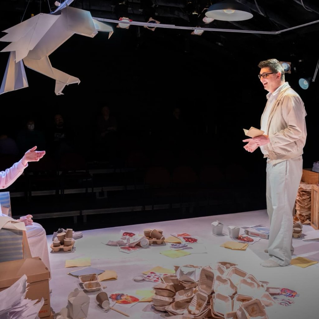 Archive photo from Animals Out of Paper. A woman is seated on a bench with a paper hawk hanging above. Across the room is a man she is talking to.