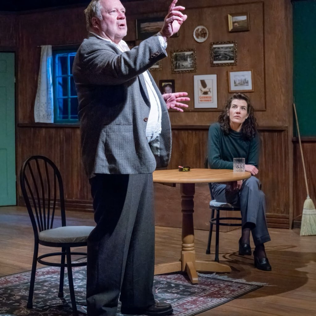 A man standing at a pub table and gesturing upwards while a seated woman looks on