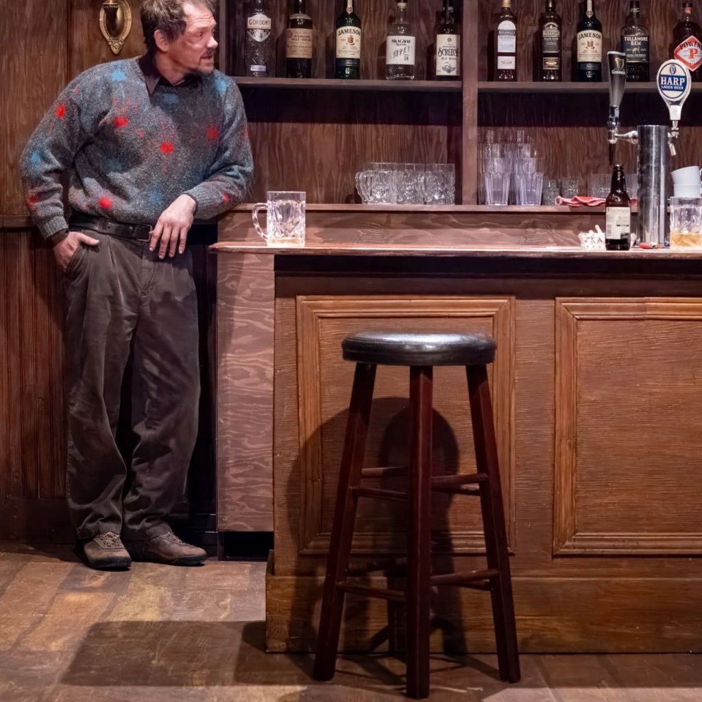 A man in a sweater leans against the liquor cabinet behind the bar in an Irish pub