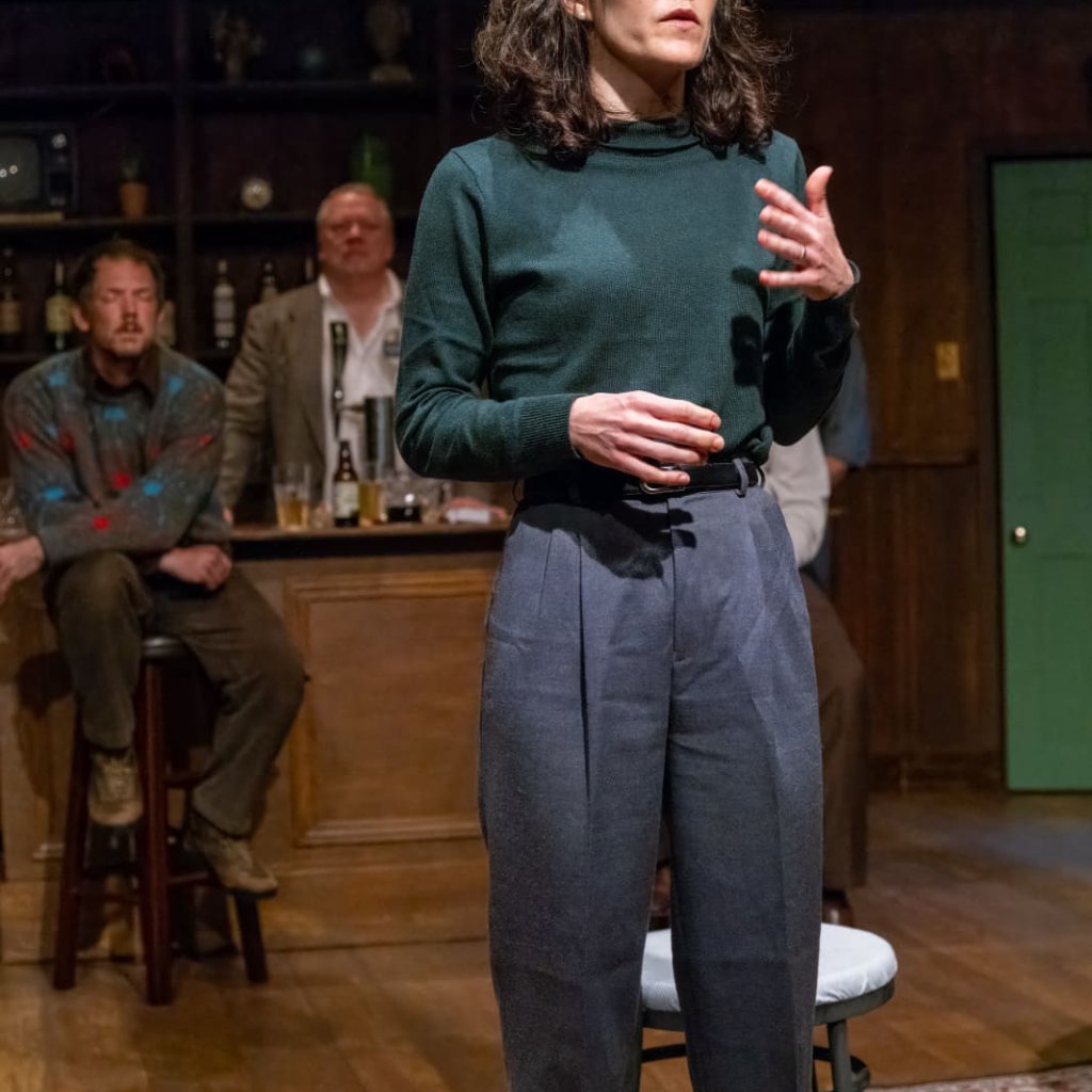 A woman in a green shirt stands and speaks, two men behind her in a dimly lit background