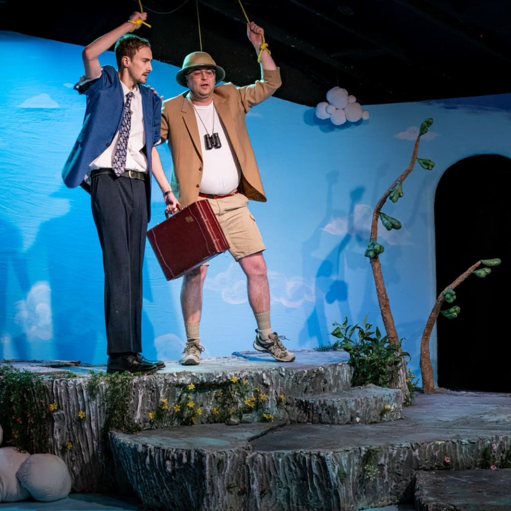 A man in a blue suit with a briefcase, and a man with a safari hat and tan blazer. They each have an arm in the air, holding a rope looped around their wrist