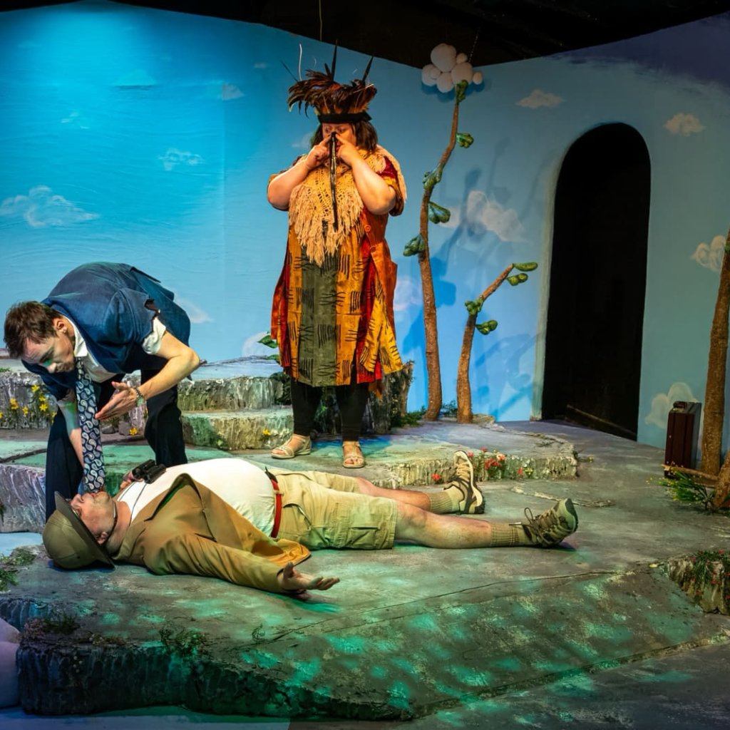 A man lies on the ground while another man leans over to speak to him. A woman dressed as a bird looks on behind them.