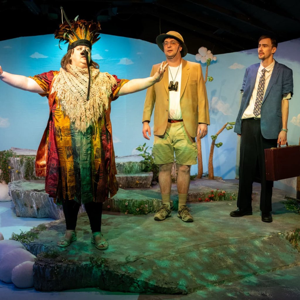 A woman dressed as a bird spreads her arms and speaks in front of two men.