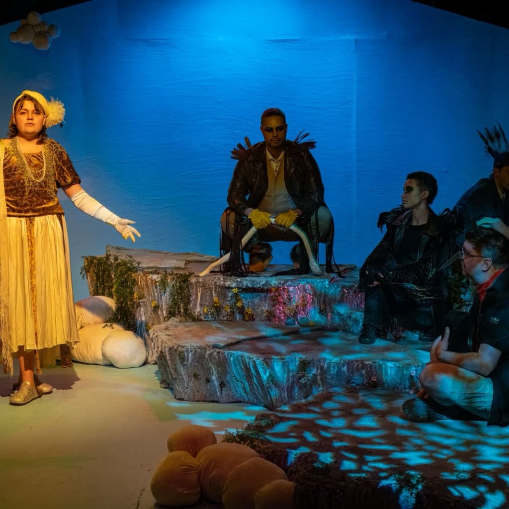 A person dressed as a nightingale sings while four others dressed like birds sit in darkness
