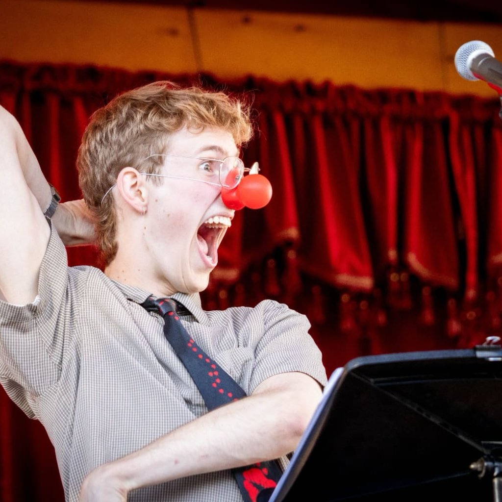 Archive photo of a Round Back performance. A young man wearing a clown nose makes a large gesture with his arms.