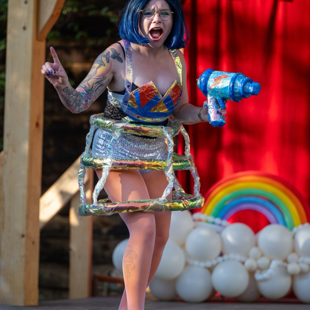 Archive photo of a Round Back performance. A performer in a colourful costume and wig with a large ray gun prop.